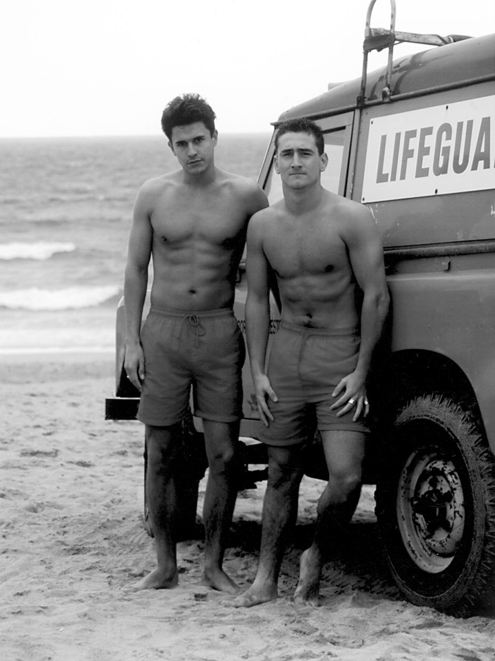 Black and white photo of two shirtless lifeguards standing by a lifeguard truck on a beach.