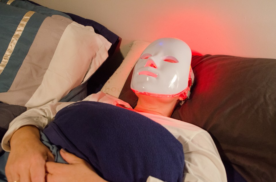 Woman using red LED light therapy mask in bed.