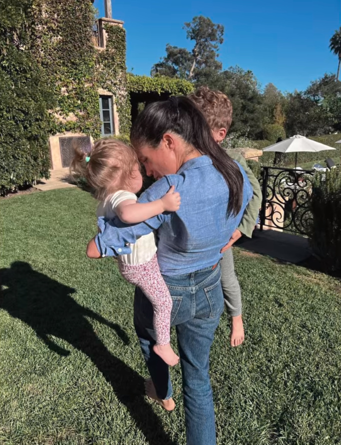 Meghan Markle carrying her children, Lilibet and Archie, on a lawn.