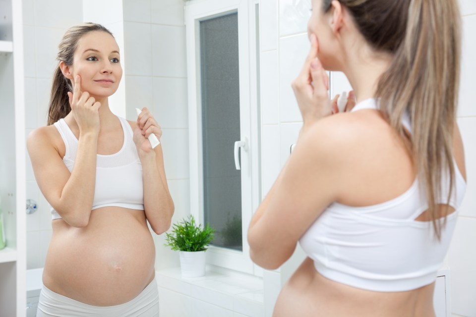 Pregnant woman applying moisturizer in front of a bathroom mirror.