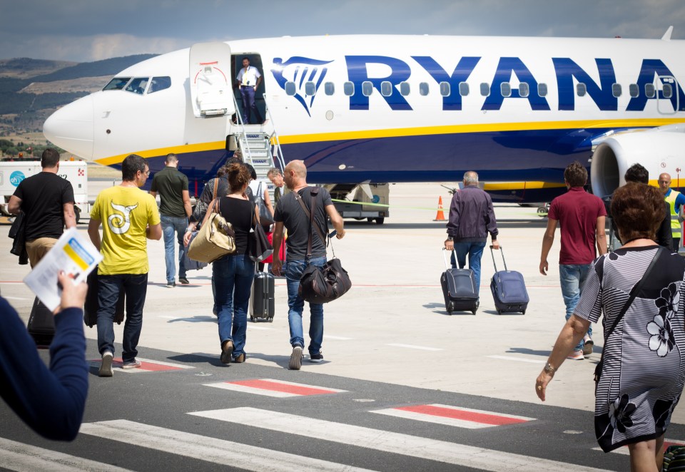 Passengers with carry-on luggage walking toward a Ryanair airplane on an airport tarmac.