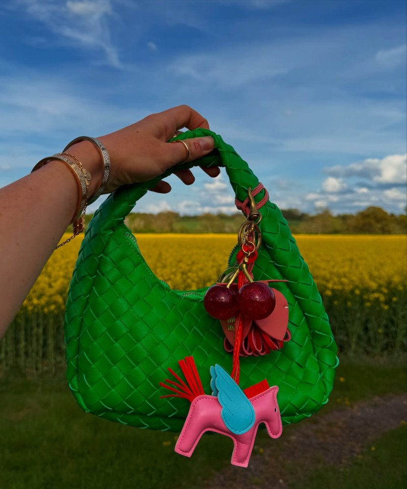 Woman holding a green woven handbag with a colorful key chain featuring cherries and a winged pony.