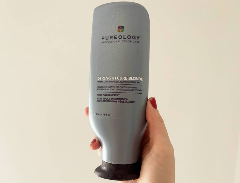 A hand holding a bottle of Pureology Strength Cure Blonde purple conditioner.