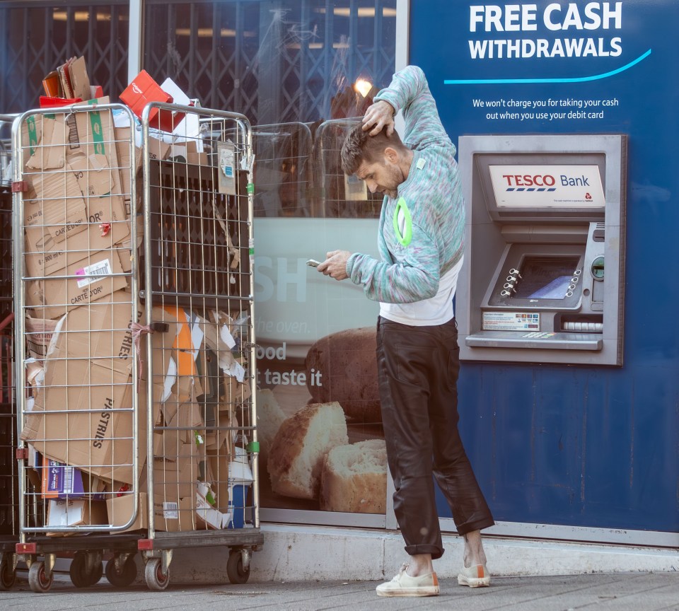 Man using phone at ATM, recycling bin nearby.