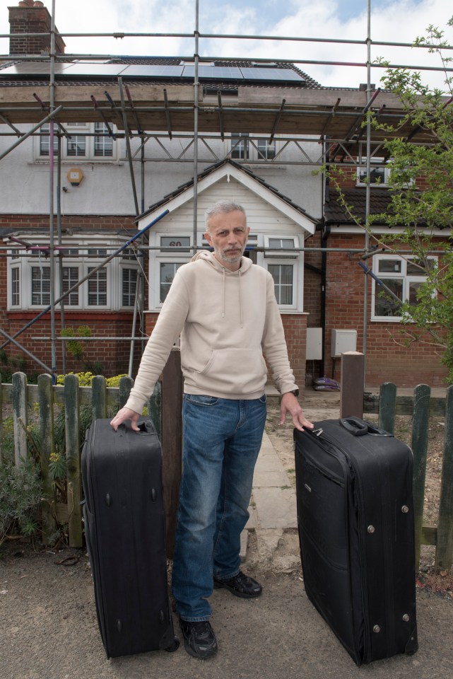A man stands outside his house under construction, holding two suitcases.