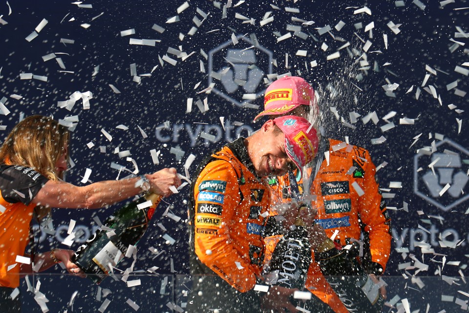 Lando Norris and Oscar Piastri celebrating on a podium with champagne and confetti.