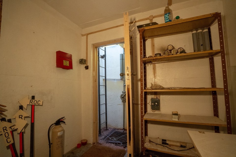 Interior of a Cold War bunker, showing a doorway with a ladder leading down, and shelves with various items.