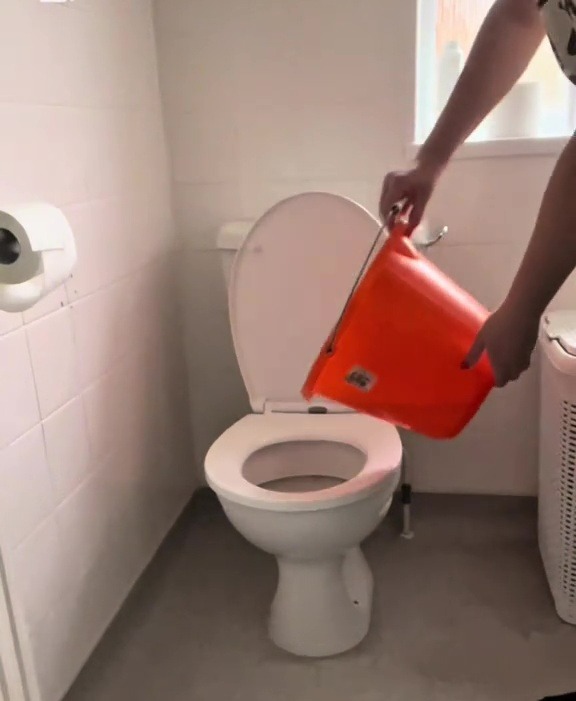Person pouring water from an orange bucket into a toilet.