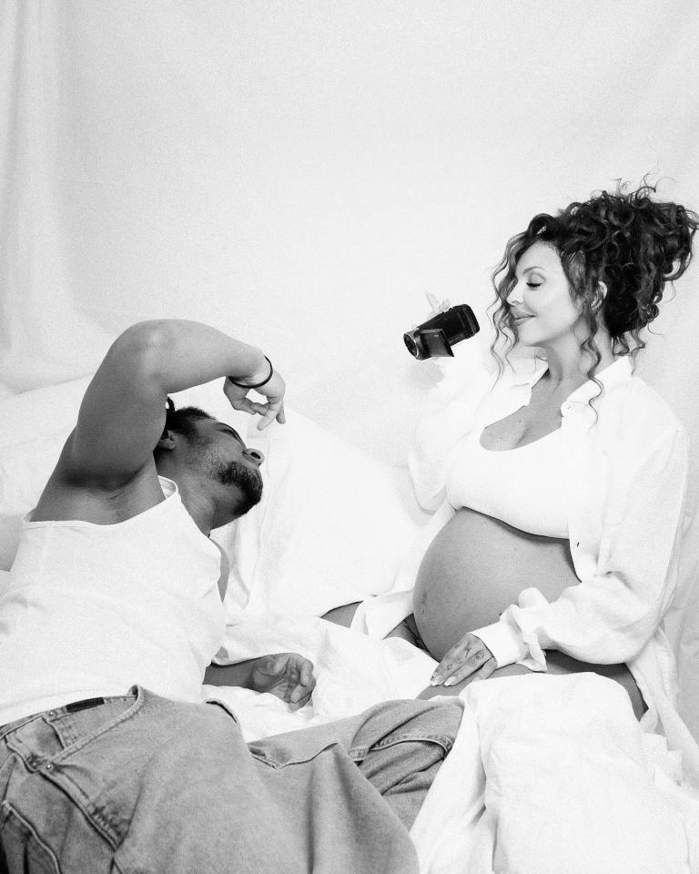 Black and white photo of a pregnant woman and a man lying in bed.  The woman holds a camcorder.