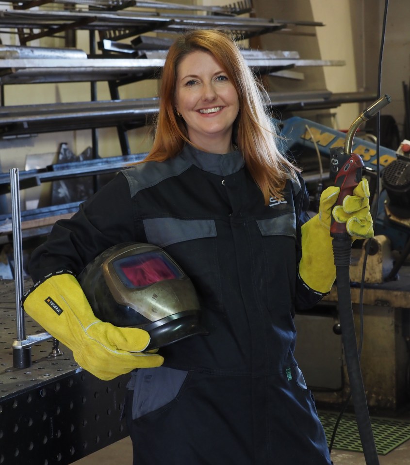 A woman in a welding suit holds a welding torch and a welding helmet.