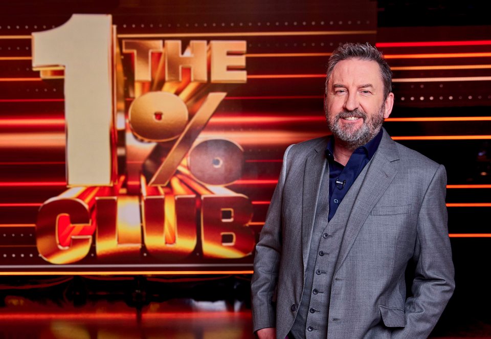 Lee Mack in a suit, standing in front of the "The 1% Club" logo.