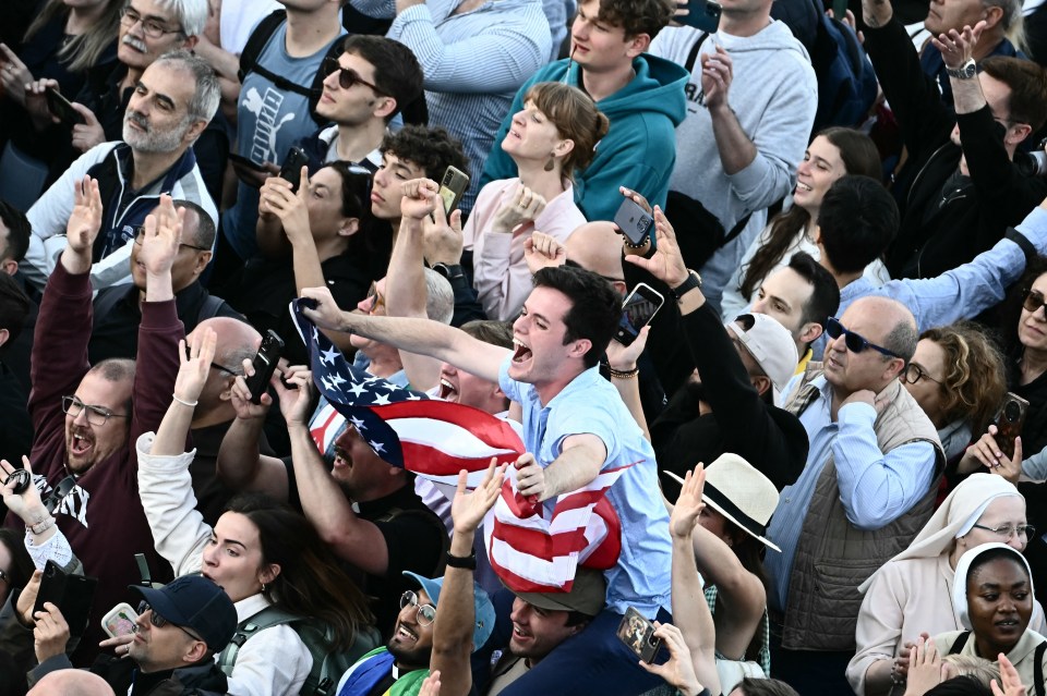 A crowd of people cheering, holding up an American flag, reacting to the announcement of a new pope.