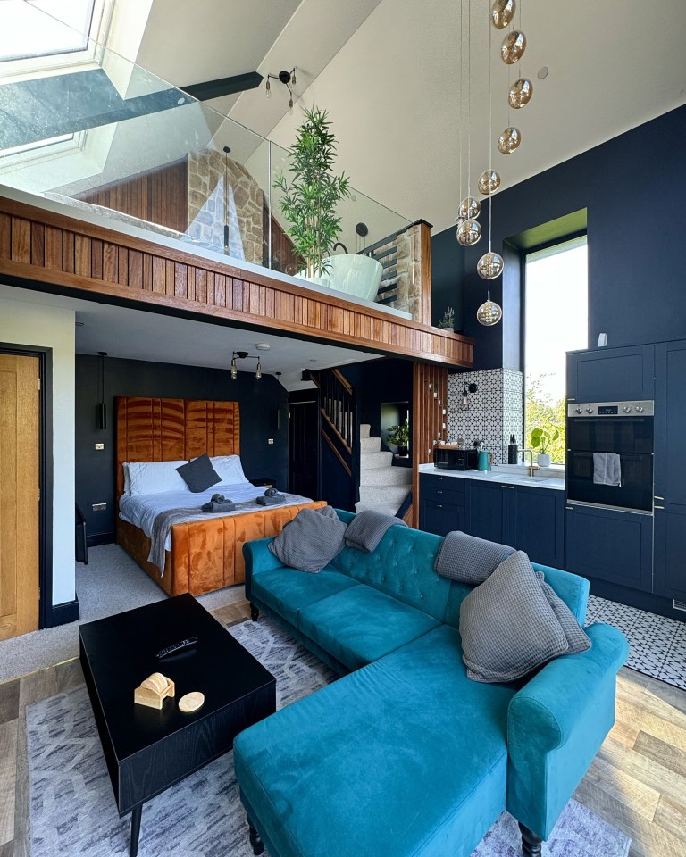 Interior view of a luxury barn featuring a teal sectional sofa, orange velvet bed, and modern kitchen.