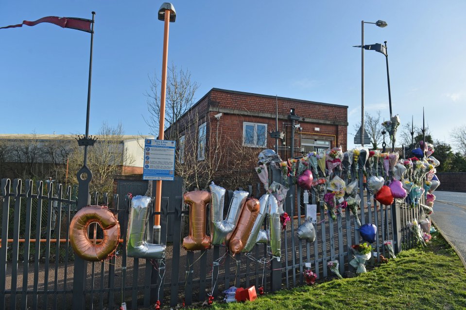 Floral tributes and balloons spelling "Olivia" attached to a fence near a railway station.