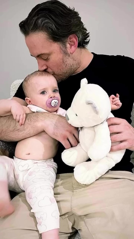 A man kisses his baby daughter, who is holding a teddy bear.