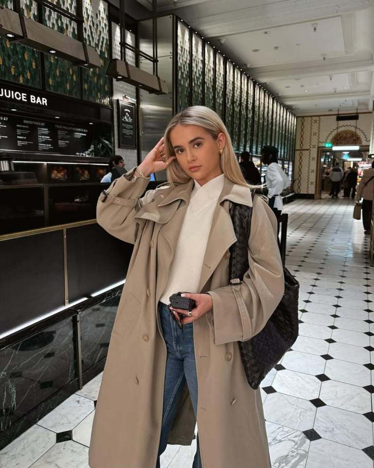 Woman in a beige trench coat and jeans in a building hallway.