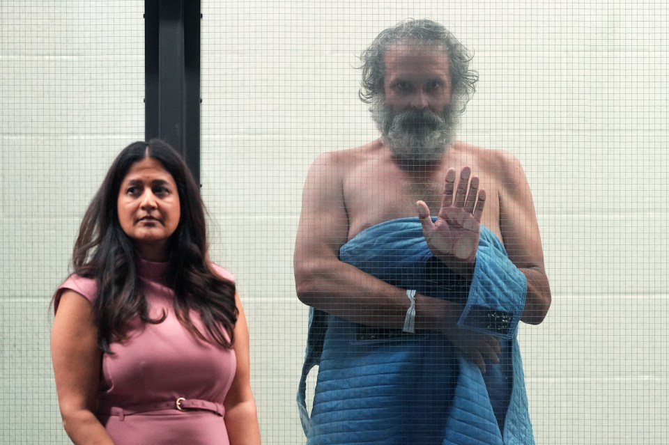 A man and woman seen through a wire mesh partition; the man is shirtless and holding up a hand.