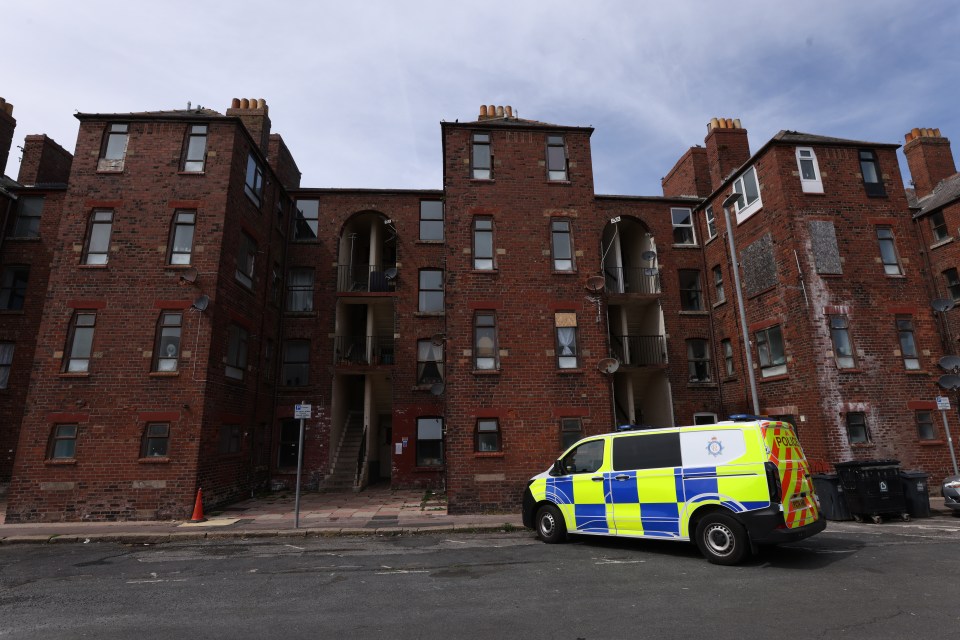 Row of brick buildings with parked cars and a police van.