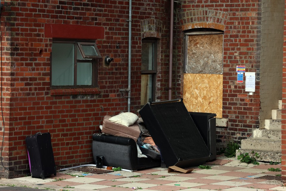 Discarded furniture and luggage outside a brick building with boarded-up windows.