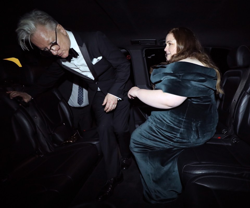 A man in a tuxedo helps a woman in a dark green velvet dress into a car.
