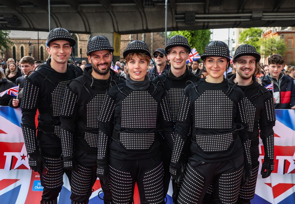 Six performers in black light-up suits on a red carpet.