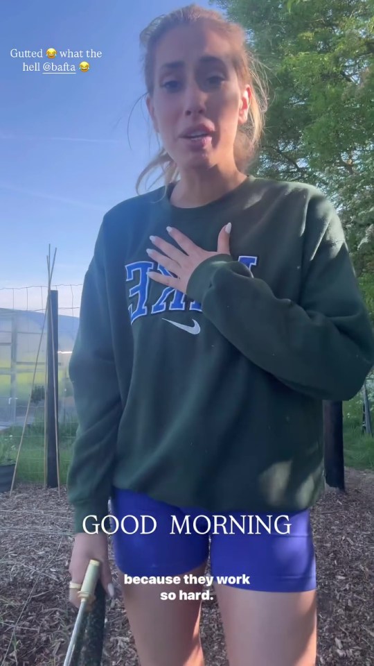 Woman in a green sweatshirt and blue shorts speaks to the camera outdoors.