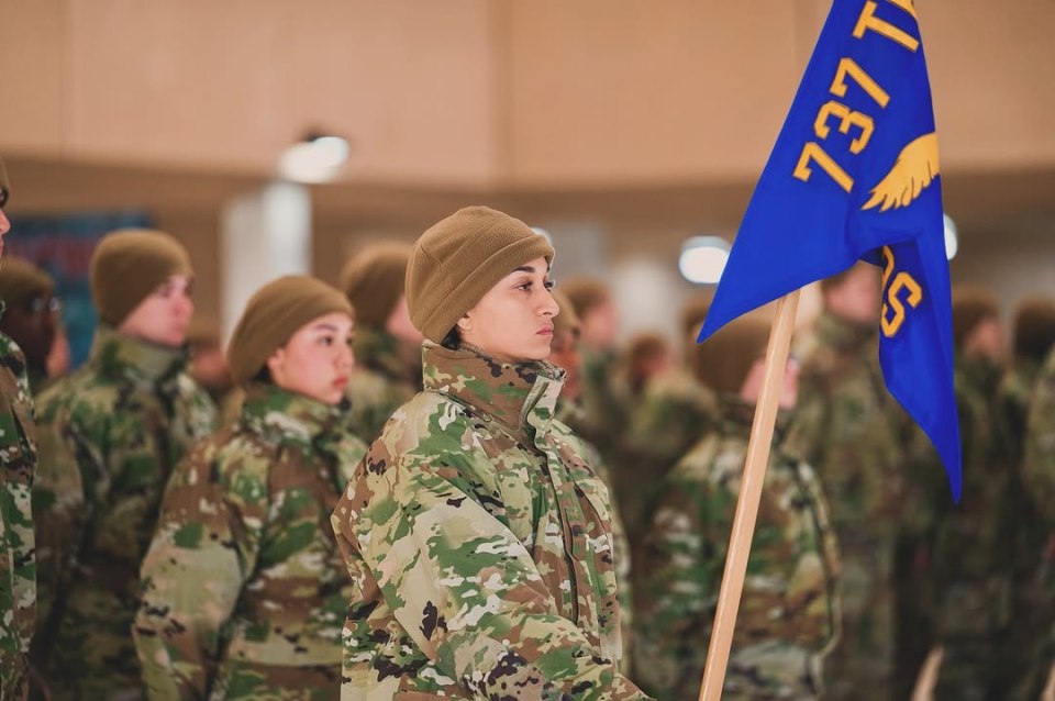A female soldier in camouflage uniform holds a blue flag with "737" on it.