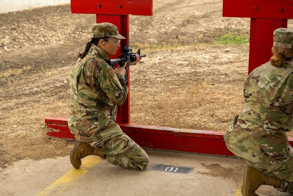 Two soldiers in camouflage kneeling at a shooting range, aiming rifles.