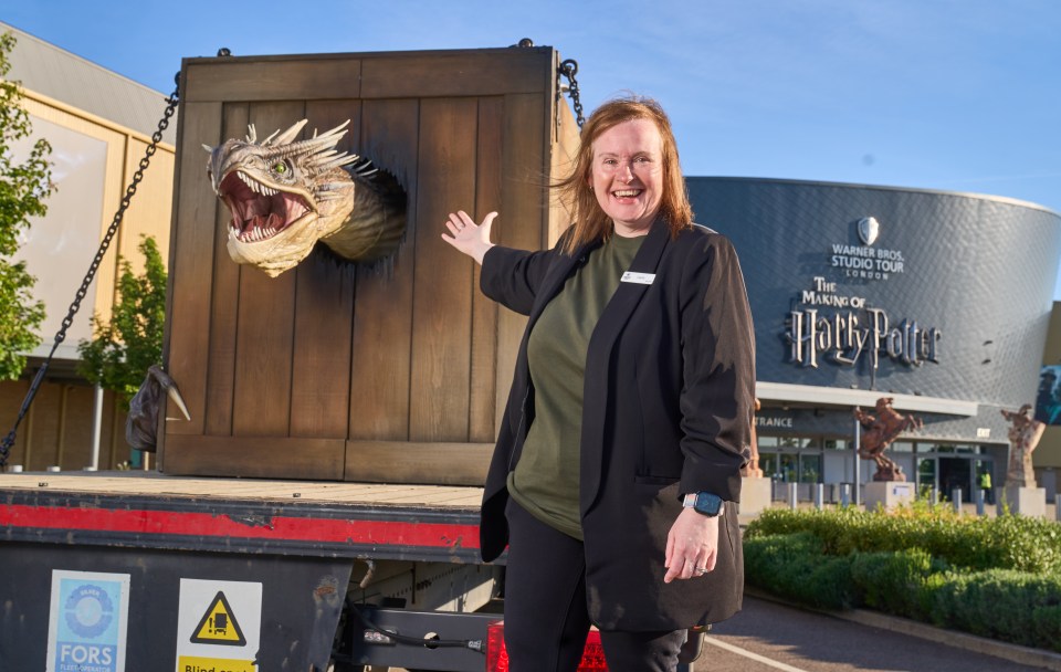 A woman stands next to a large animatronic dragon head on a truck, in front of the Warner Bros. Studio Tour London – The Making of Harry Potter.