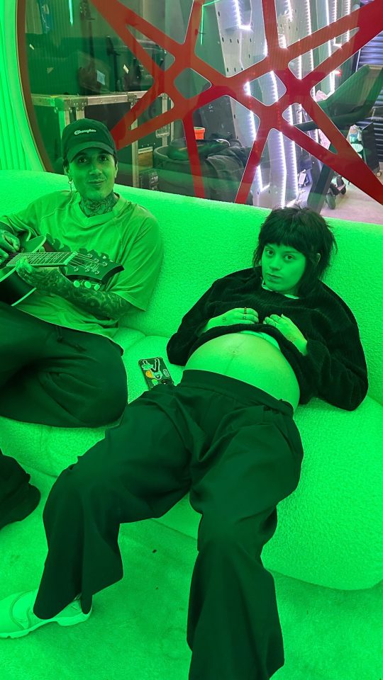 Pregnant person and man sitting on a green couch; the man is holding a guitar.