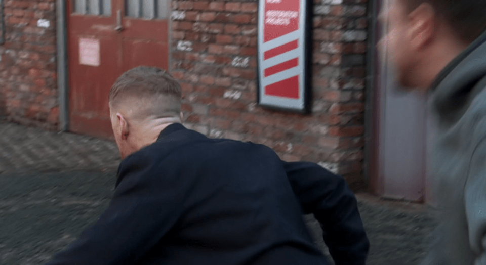 Screenshot of a man running away from another man in an alleyway.