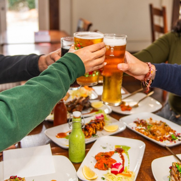 People toasting with beer glasses at a restaurant table with Indian food.