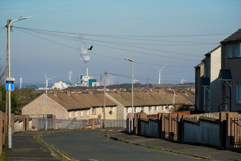 Street scene in Workington, England, showing terraced houses, a factory in the distance, and wind turbines on the horizon.