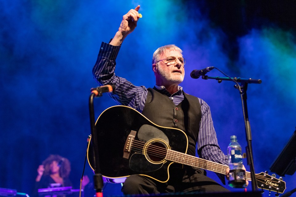 Steve Harley performing on stage with an acoustic guitar.