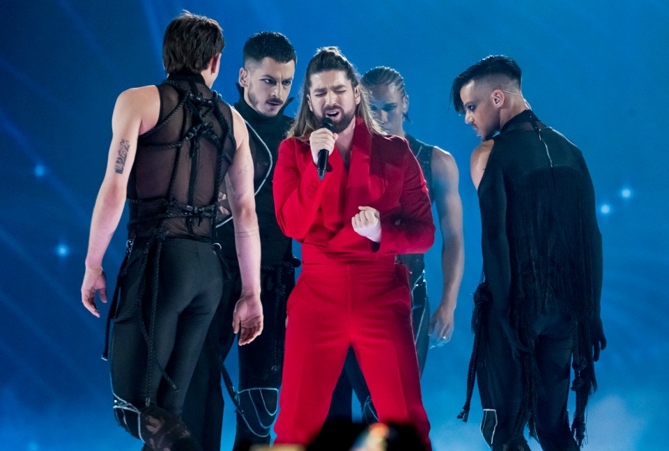 A singer in a red suit performs on stage with backup dancers.