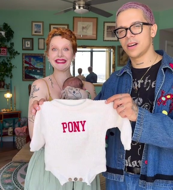 Parents holding their newborn baby, who is wearing a white sweater that says "PONY" on it.
