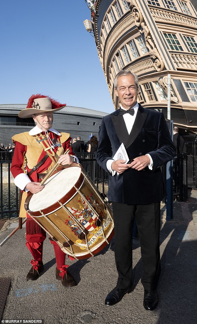 A beaming Nigel Farage donning a dark velvet blazer coupled with a bow tie, next to a drummer. Behind him is HMS Victory, Britain's oldest commissioned warship