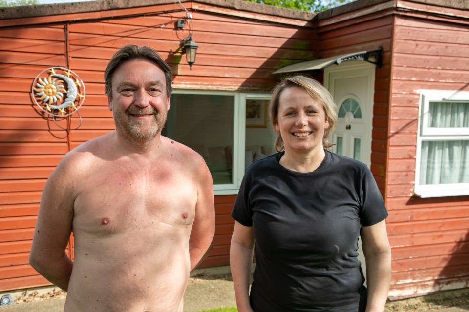 A shirtless man and a woman wearing a black t-shirt stand outside a red cabin.