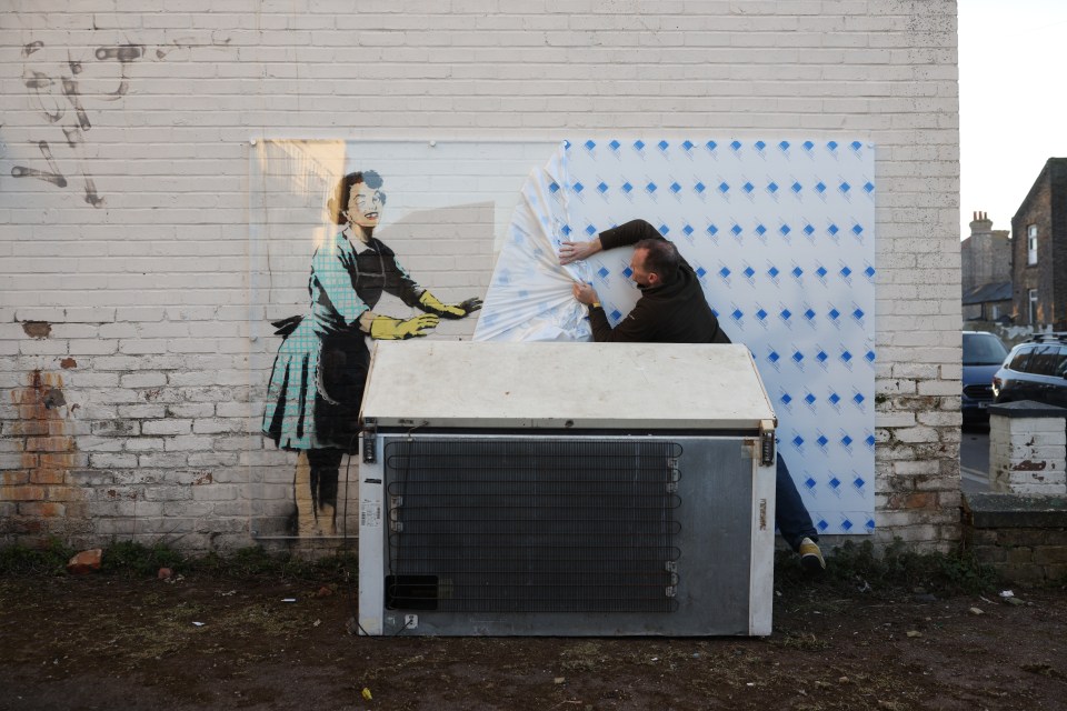 A worker covering a Banksy artwork depicting a woman in a 1950s-style dress with a protective screen.