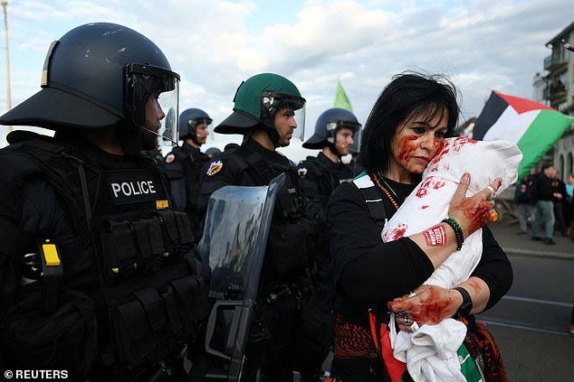 A woman holding a prop representing a body bag grips it tightly to her face as armed police officers stand nearby