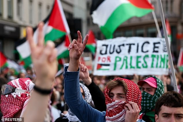 Protesters dressed in keffiyehs hold peace signs into the air, as a sign which reads 'Justice for Palestine' is waved in the background