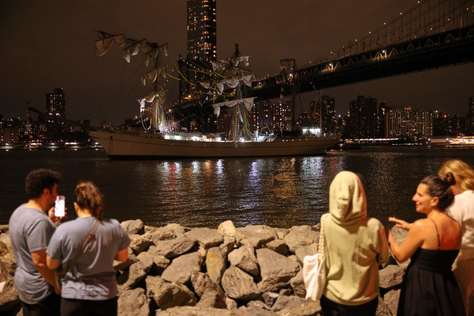 Pedestrians watching a ship under the Brooklyn Bridge at night.