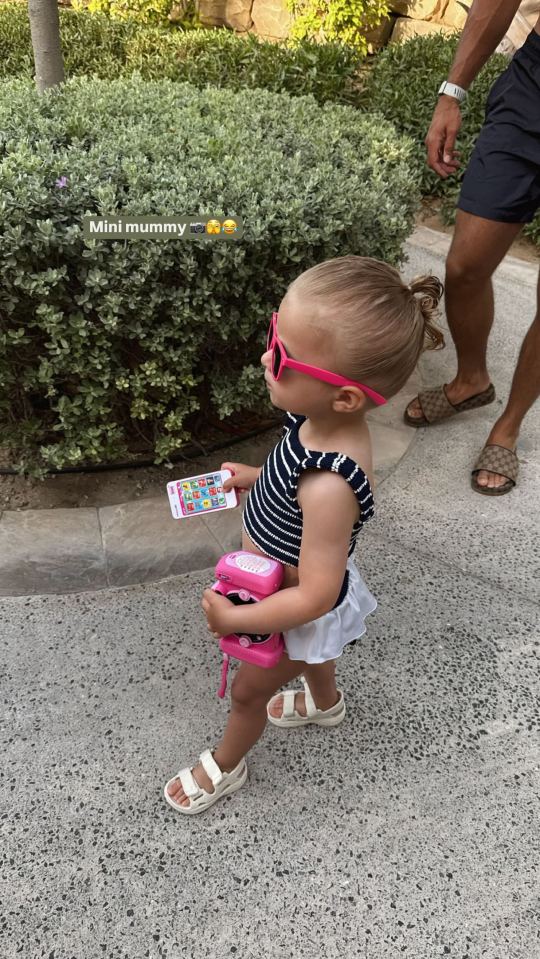 Toddler girl wearing sunglasses and holding toy phone and camera.