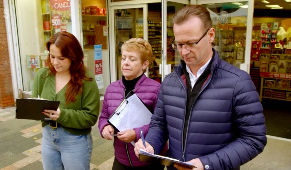 Three people filling out clipboards outside a shop.