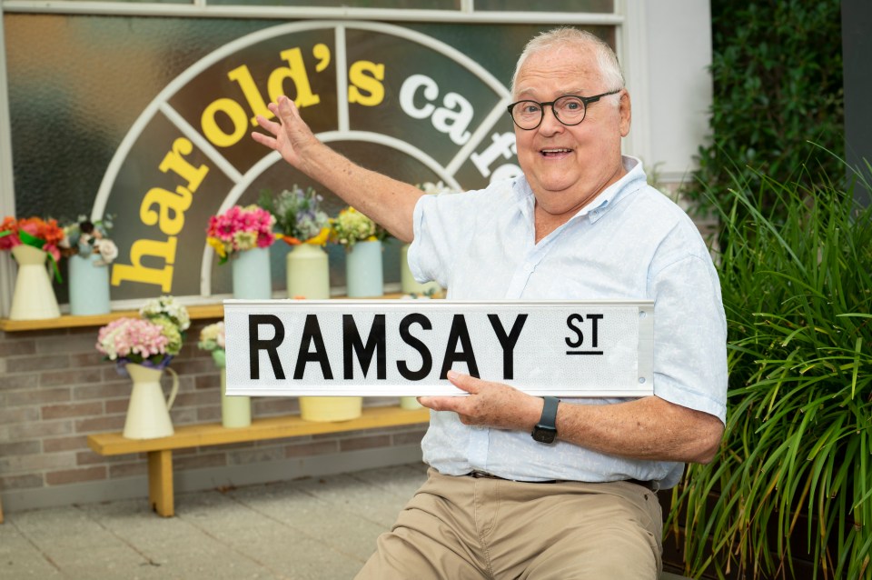 Ian Smith holding a Ramsay Street sign in front of Harold's Cafe.