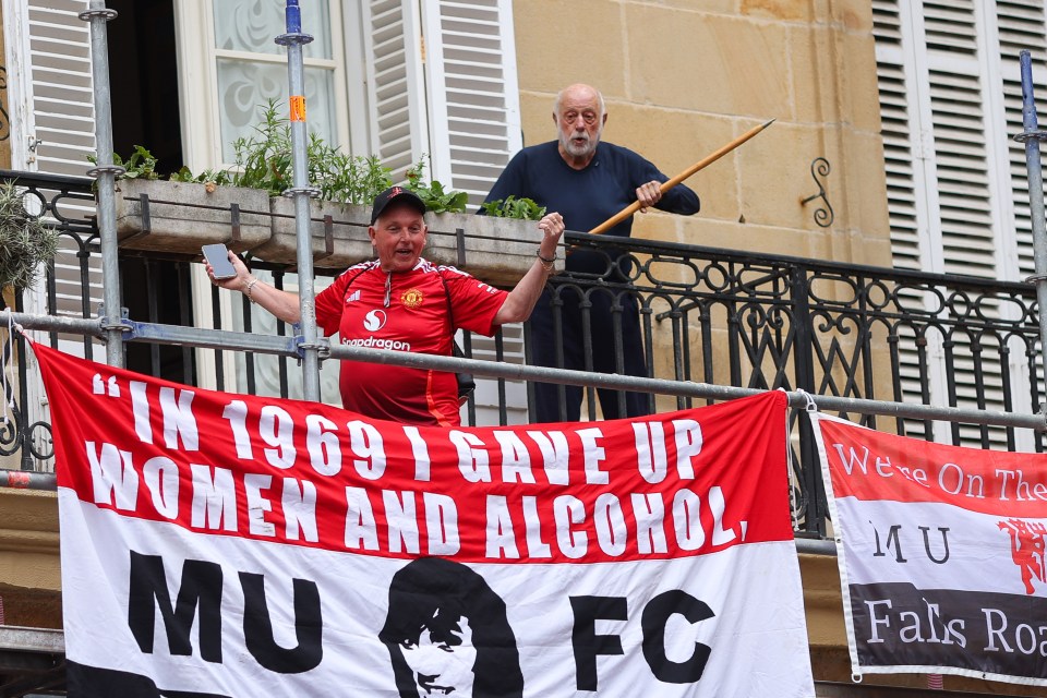 A Manchester United fan on a balcony holds a banner that reads, "In 1969 I gave up women and alcohol," while another person on the balcony holds a stick.