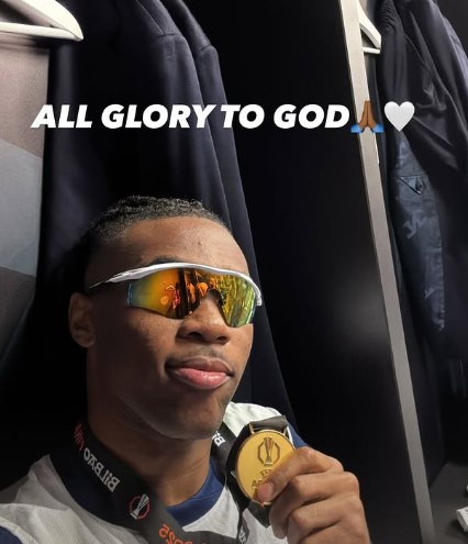 A man in sunglasses holding a gold medal in a dressing room.