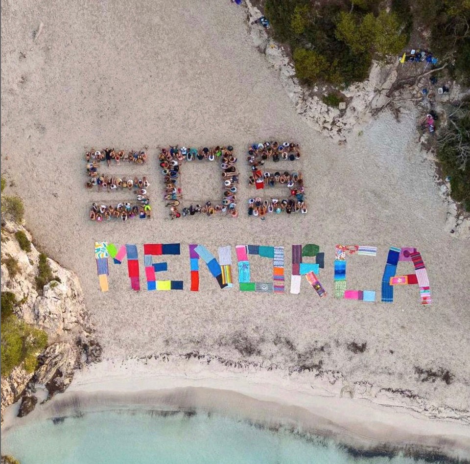 Aerial view of protestors on a beach forming the letters SOS MENORCA with their bodies and towels.
