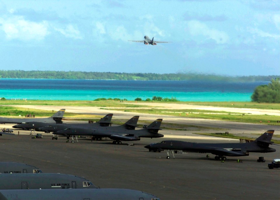 Photo of B-1 bombers on a runway with a B-1 taking off in the background, overlooking a tropical atoll.