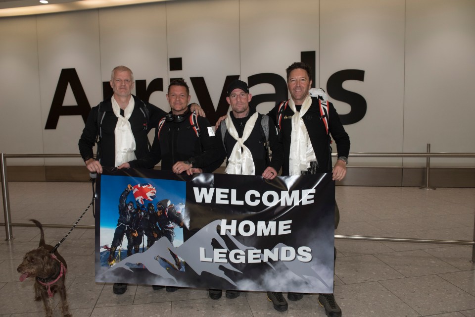 Four men at an airport arrivals gate holding a banner that says "Welcome Home Legends," with a photo of them on a mountaintop.